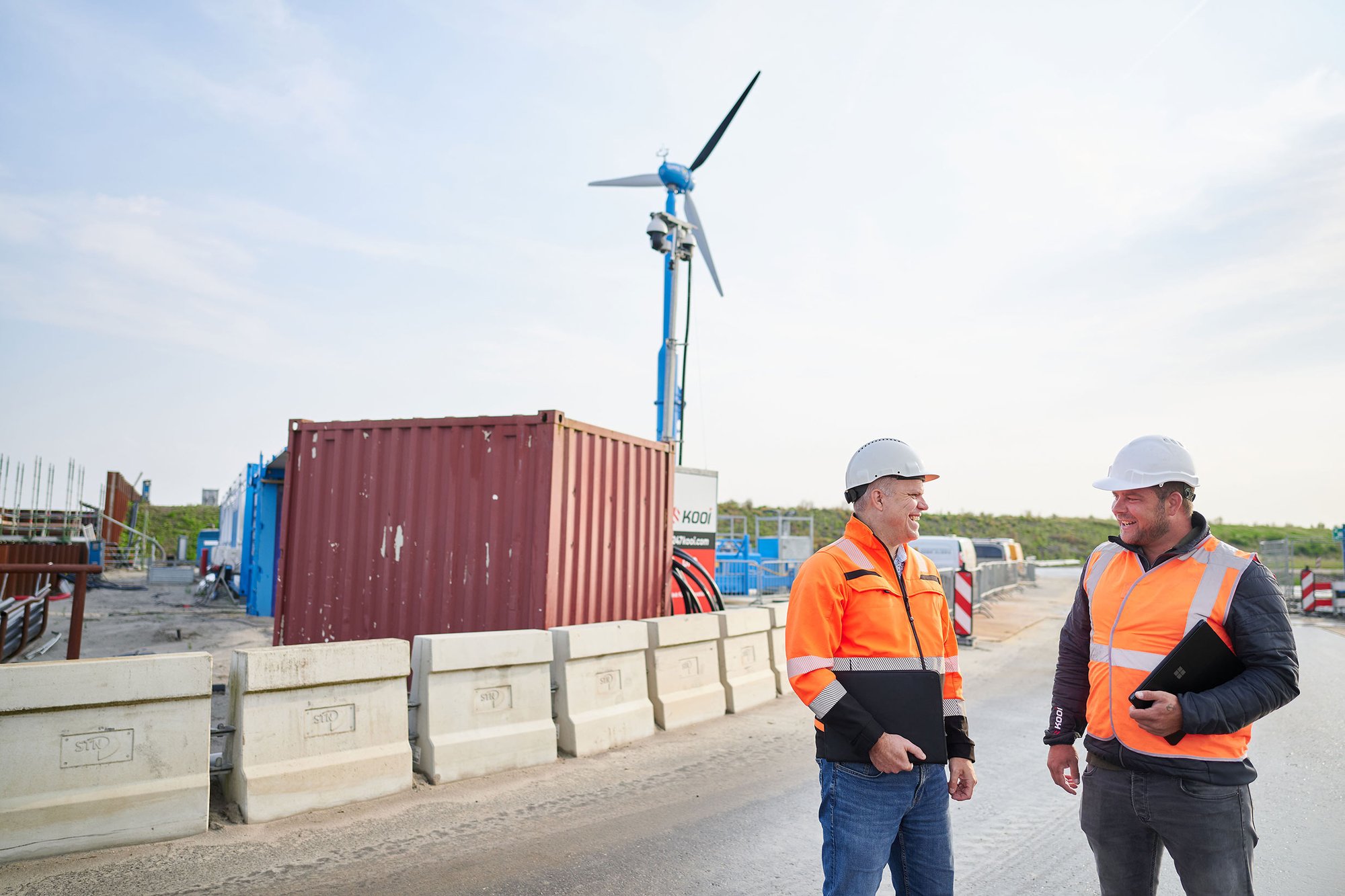 Two security professionals in high-visibility gear and helmets discussing site operations near a Kooi surveillance system at a construction site with a wind turbine in the background, highlighting safety and monitoring in sustainable environments.