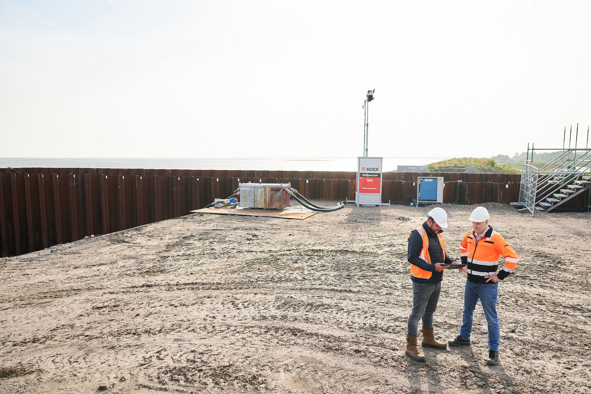 Two security professionals in high-visibility gear and helmets discussing operations at a monitored construction site with a Kooi surveillance system, highlighting safety and reliable site management.