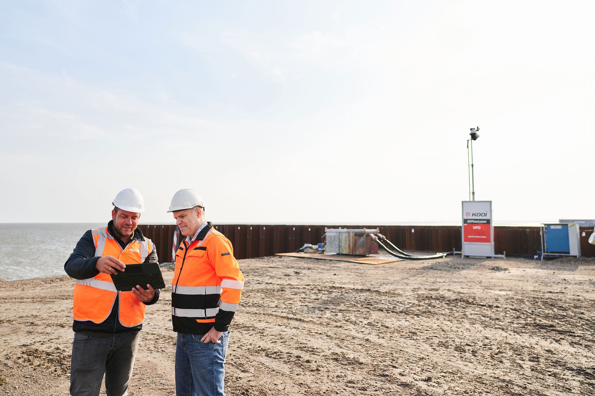 Two security professionals in high-visibility jackets and helmets reviewing site plans on a tablet at a monitored waterfront construction site, showcasing effective collaboration and reliable monitoring with Kooi surveillance.