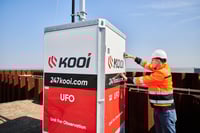 Construction professional in high-visibility gear and a helmet inspecting a Kooi surveillance unit at a construction site, showcasing advanced monitoring technology and proactive site management.