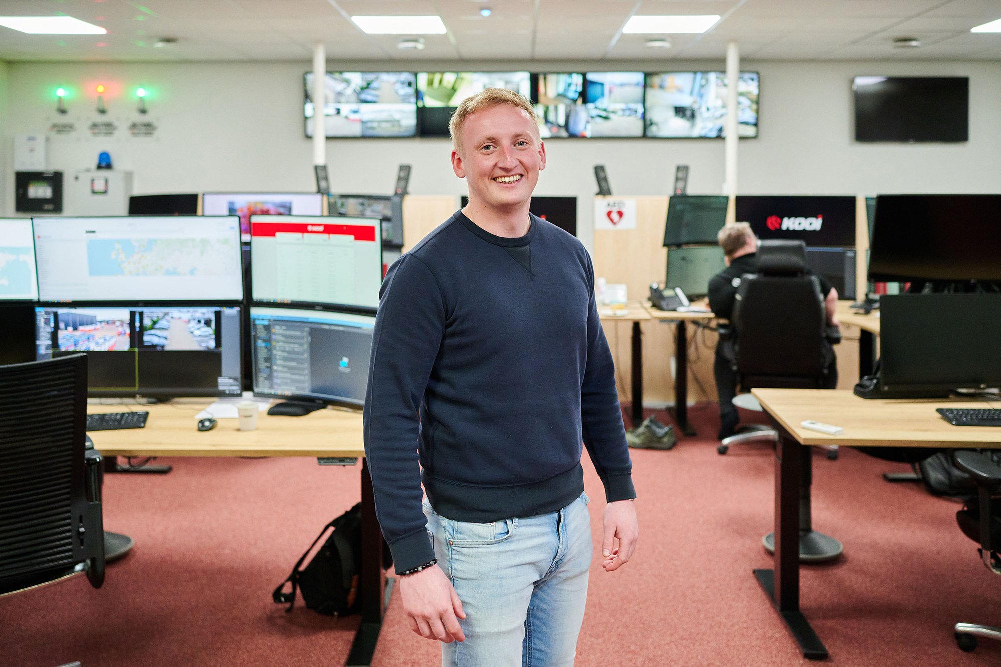 Kooi employee standing confidently in a control room with multiple monitoring screens, showcasing a reliable, innovative, and technology-focused approach to security operations.