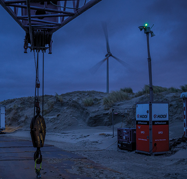 Kooi security units monitoring a construction site at night, with a wind turbine and equipment in the background, emphasizing a reliable, innovative, and robust approach to site safety and security.