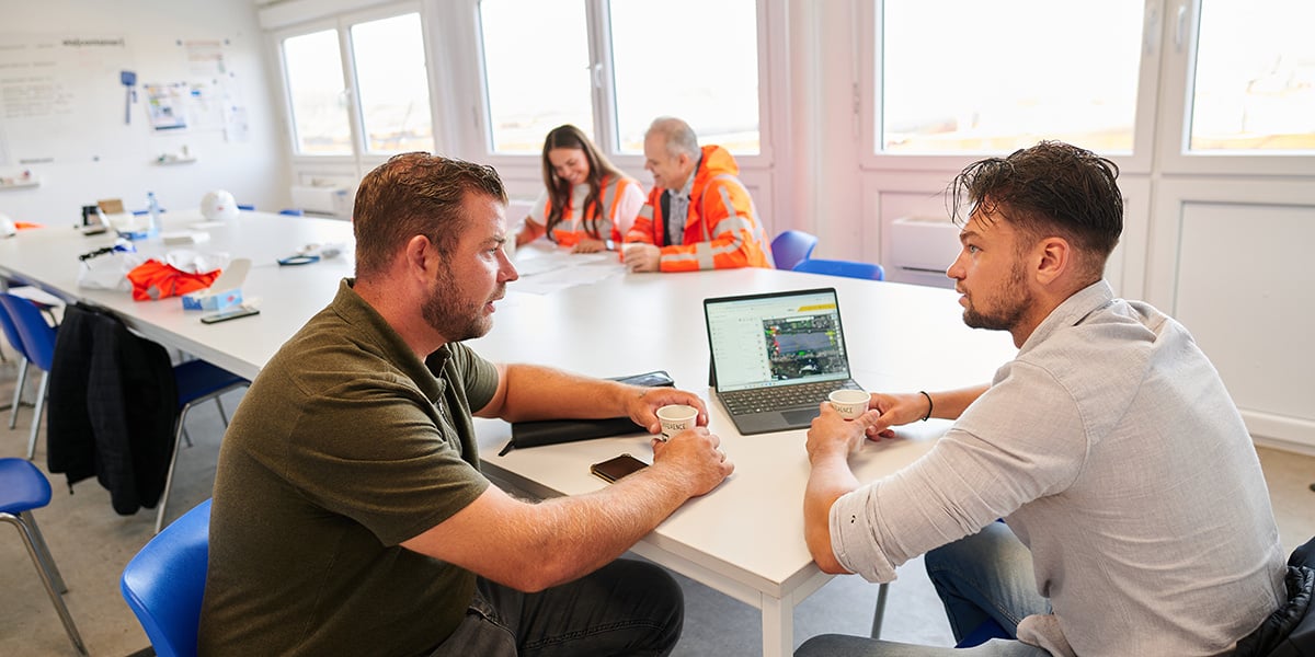 Kooi employees discussing project details over coffee with a laptop on the table, while colleagues collaborate in the background, showcasing a reliable, innovative, and teamwork-driven work environment.
