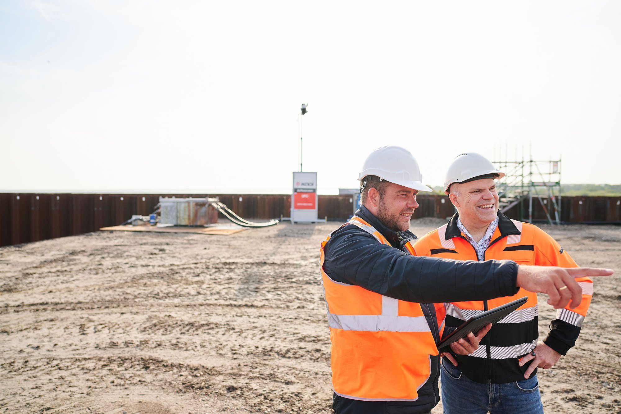 Due professionisti della sicurezza con giubbotti ad alta visibilità e caschi discutono e indicano le attività in un cantiere monitorato, con un sistema di sorveglianza Kooi sullo sfondo, dimostrando una gestione efficace del sito e la sicurezza.