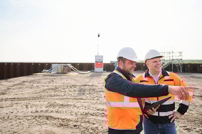 Deux professionnels de la sécurité, vêtus de vestes haute visibilité et de casques, discutant et pointant du doigt les activités sur un chantier surveillé, avec un système de surveillance Kooi en arrière-plan, illustrant une gestion et une sécurité de site efficaces.